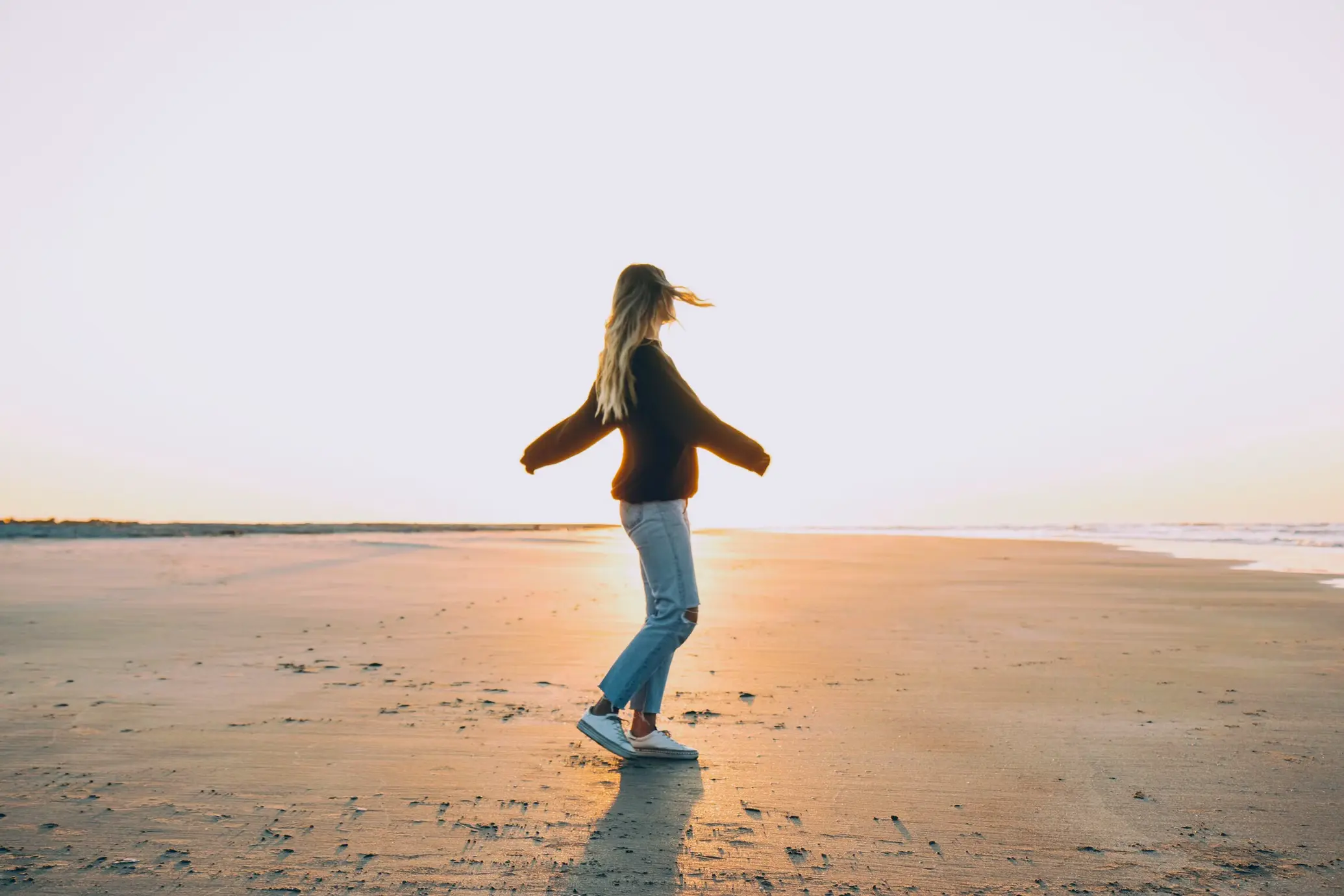 A woman with long blonde hair spinning on a sandy beach during sunset, wearing a dark sweater and light-colored jeans.