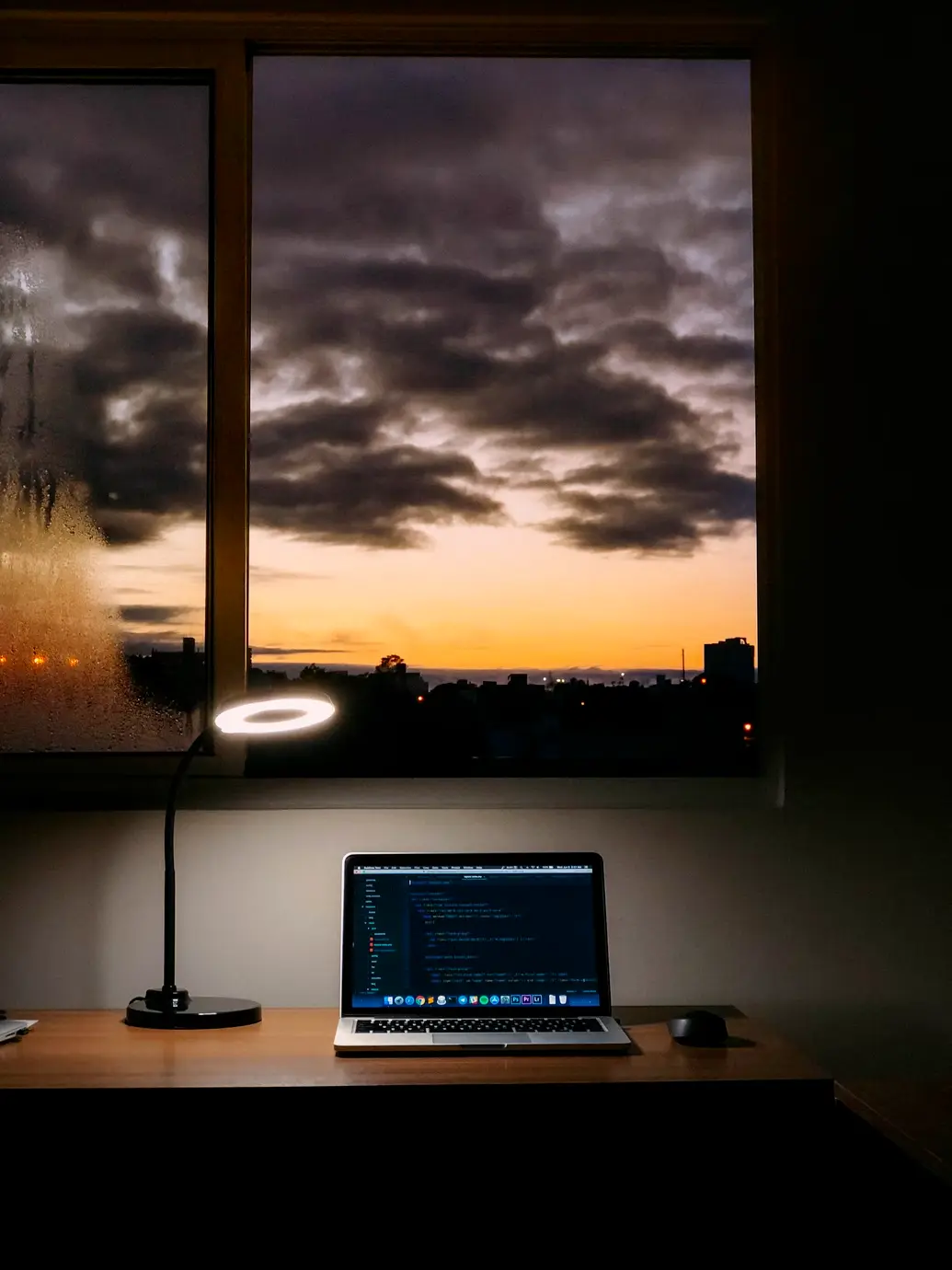 A laptop with a code editor on a desk under a ring lamp, with a city sunset visible through the window.